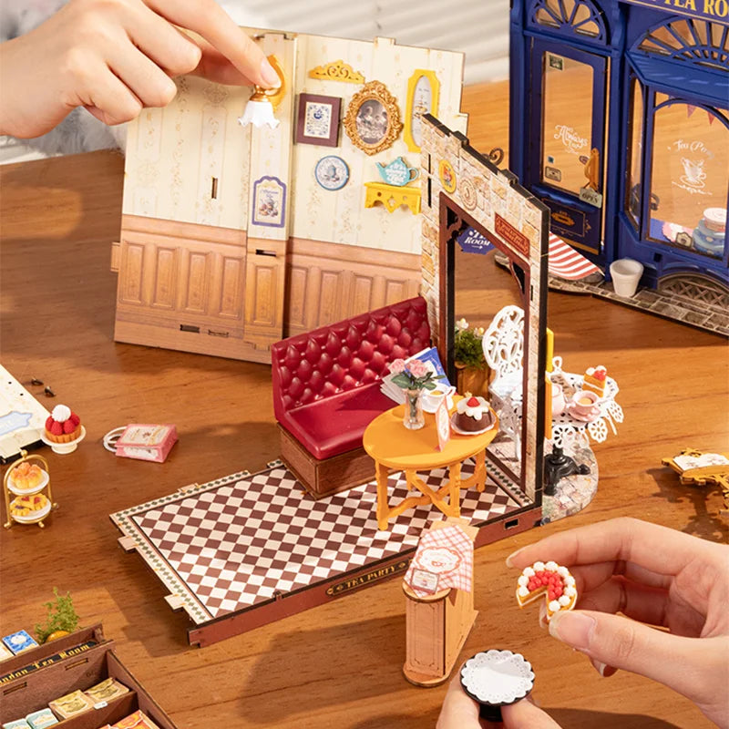 Hands assembling miniature DIY dollhouse tea shop kit with tiny furniture and pastries.